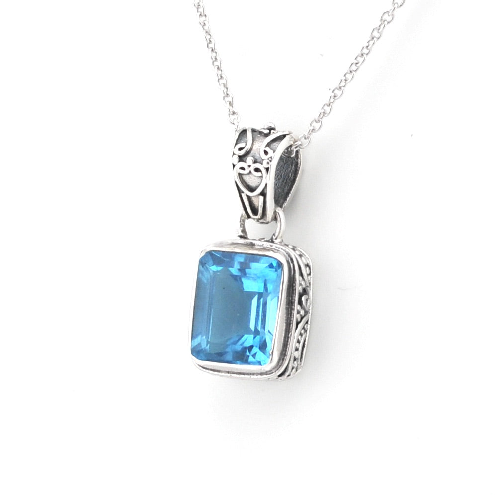 A silver pendant necklace featuring a rectangular, emerald-cut blue gemstone in an ornate silver bezel setting, suspended from a delicate silver chain against a plain white background.