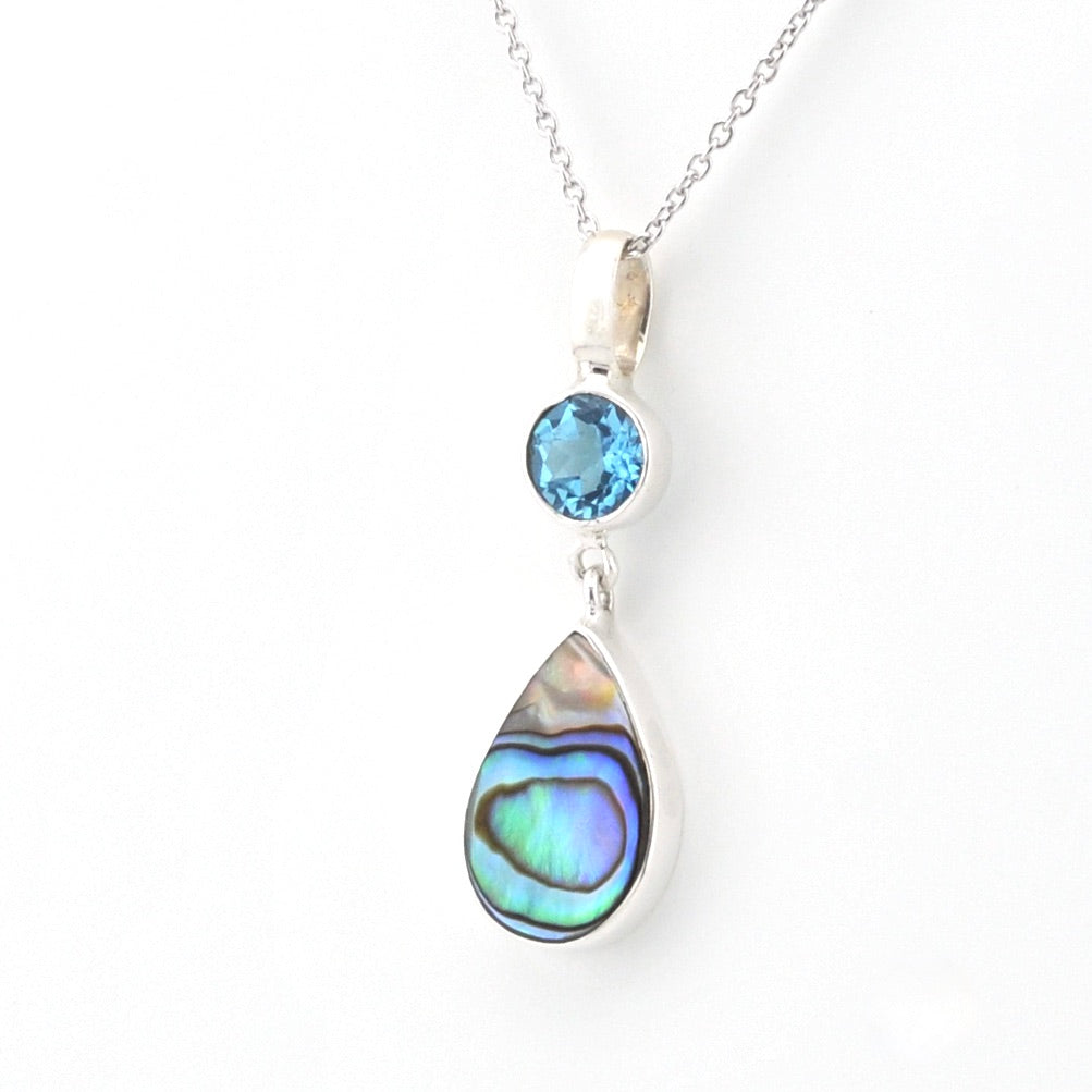 A silver pendant necklace featuring a round light blue gemstone and a larger teardrop-shaped abalone shell inlay with iridescent blue, green, and purple hues, shown on a white background.