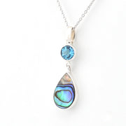 A silver pendant necklace featuring a round light blue gemstone and a larger teardrop-shaped abalone shell inlay with iridescent blue, green, and purple hues, shown on a white background.