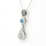 A silver-toned dangle pendant necklace featuring a small square blue gemstone and a larger teardrop-shaped milky white gemstone, suspended from a delicate silver chain against a plain white background.