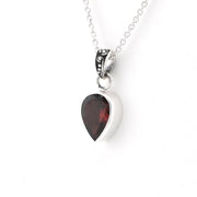 A silver heart pendant necklace featuring a faceted dark red gemstone, suspended from a delicate silver chain against a plain white background.
