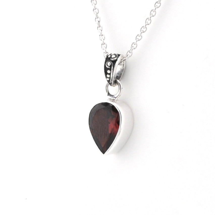A silver heart pendant necklace featuring a faceted dark red gemstone, suspended from a delicate silver chain against a plain white background.