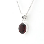 A silver pendant necklace featuring an oval dark red garnet gemstone in a polished bezel setting, suspended from a delicate silver chain against a plain white background.