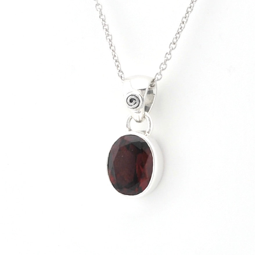 A silver pendant necklace featuring an oval dark red garnet gemstone in a polished bezel setting, suspended from a delicate silver chain against a plain white background.