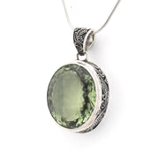 An oval pendant necklace featuring a faceted olive-green gemstone in a silver bezel with ornate filigree detailing on the bail and sides, suspended from a silver chain against a plain white background.