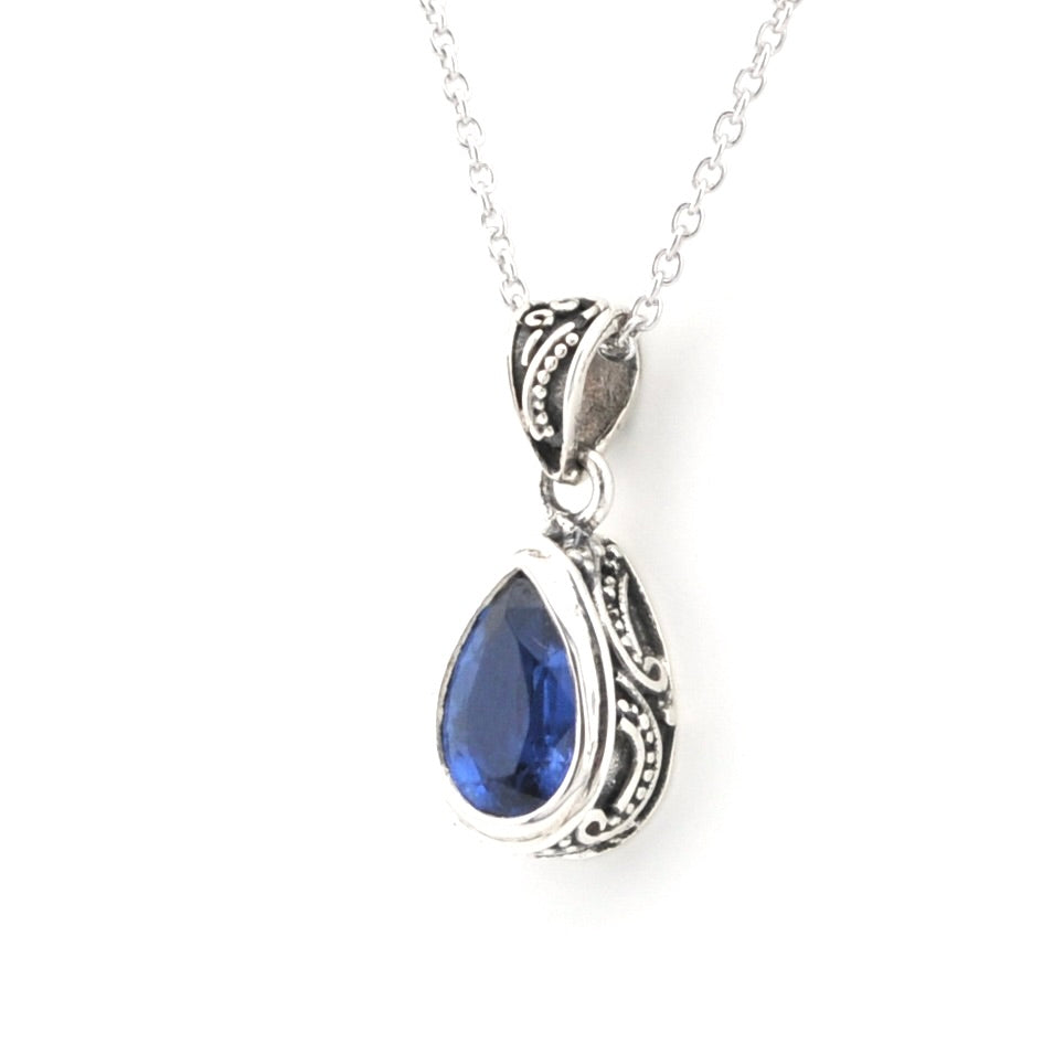 A silver teardrop pendant necklace featuring a deep blue gemstone in an ornate filigree setting, suspended from a delicate silver chain against a white background.