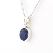 A silver pendant necklace featuring an oval-cut deep blue gemstone in a polished silver bezel setting, suspended from a delicate silver chain against a plain white background.