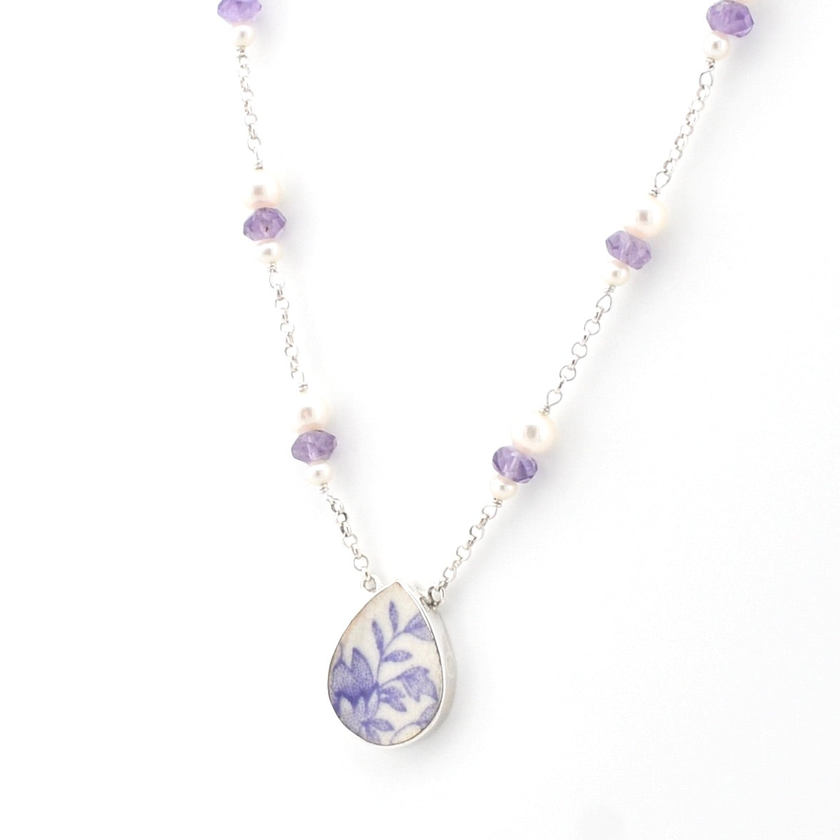 A silver chain necklace featuring a teardrop-shaped pendant with a blue floral pattern on a white background, accented by small white pearls and purple beads on a white studio background.