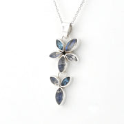 A silver pendant necklace featuring a vertical arrangement of marquise-cut blue and smoky gray gemstones in a floral leaf design, suspended from a delicate silver chain against a plain white background.