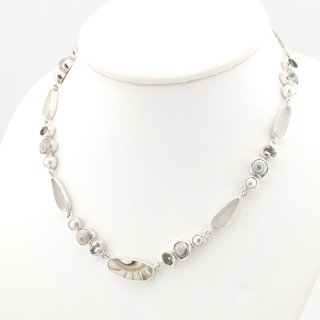 A silver-toned beaded necklace displayed on a white mannequin bust. The necklace features a mix of polished shell, white topaz, small white pearl, and gray stone.