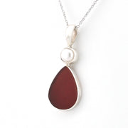 A silver pendant necklace featuring a teardrop-shaped dark red sea glass stone and a small white pearl set in a silver bezel, suspended from a silver chain on a white background.