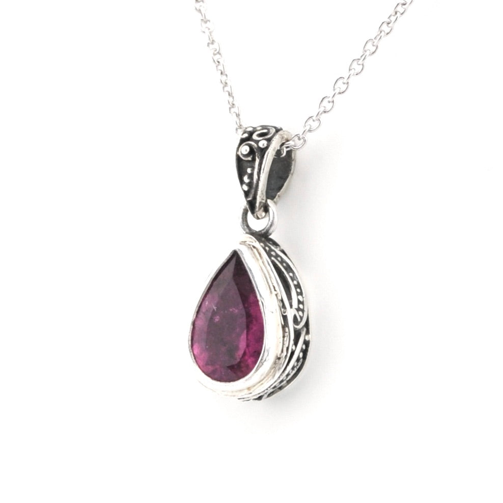A silver teardrop pendant necklace featuring a faceted dark pinkish-red gemstone, set in an ornate silver bezel with a delicate silver chain against a white background.