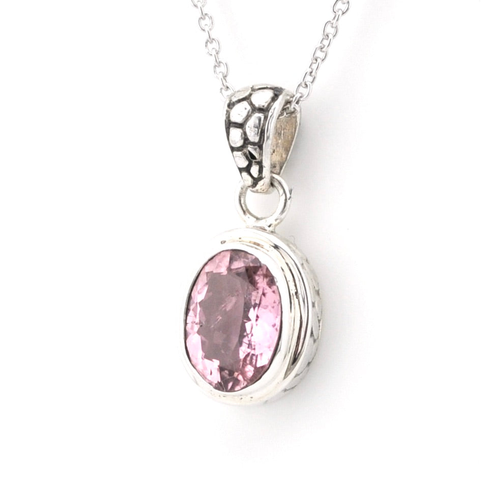 A silver pendant necklace featuring an oval-cut pink gemstone in a textured bezel setting, suspended from a delicate silver chain against a plain white background.