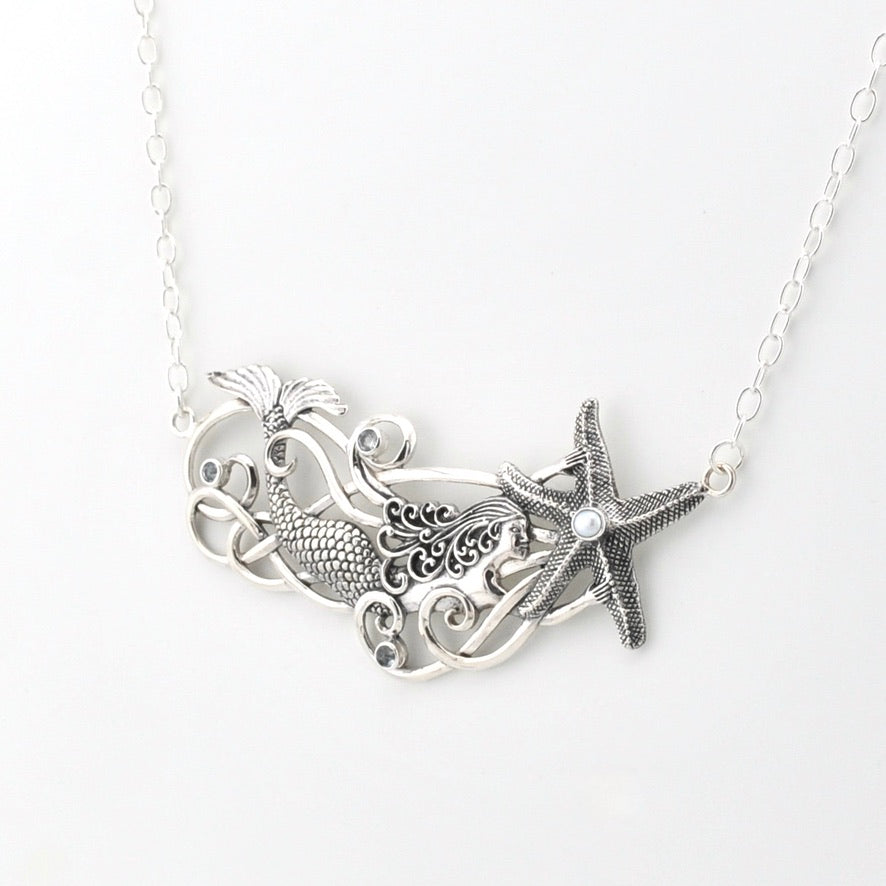 A silver-toned necklace featuring a mermaid and starfish pendant design, suspended from a delicate link chain against a plain white background.