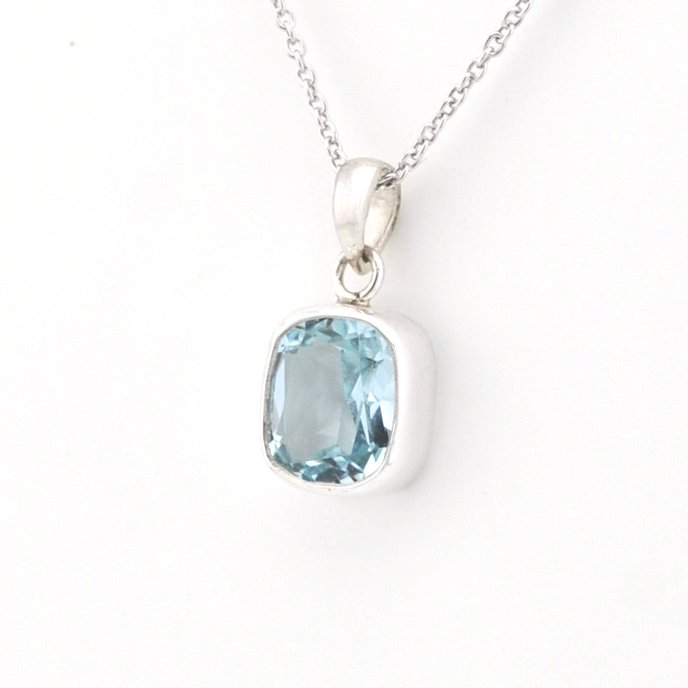 A silver pendant necklace featuring a cushion-cut light blue gemstone in a bezel setting, suspended from a delicate silver chain against a plain white background.