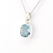 A silver pendant necklace featuring a cushion-cut light blue gemstone in a bezel setting, suspended from a delicate silver chain against a plain white background.