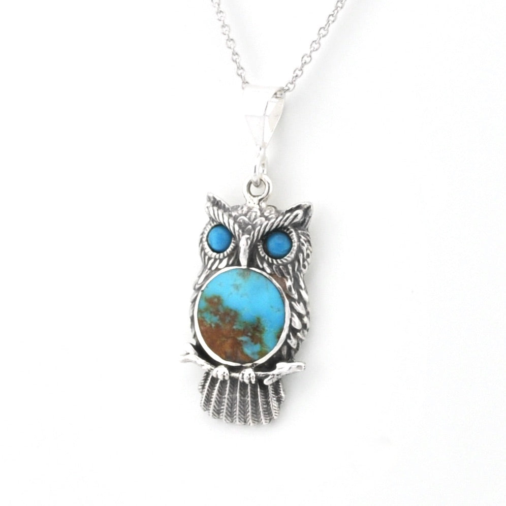 An owl pendant necklace featuring a textured silver owl body with a large oval turquoise stone in the center and two smaller round turquoise stones for the eyes, suspended from a silver chain against a white background.