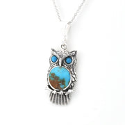 An owl pendant necklace featuring a textured silver owl body with a large oval turquoise stone in the center and two smaller round turquoise stones for the eyes, suspended from a silver chain against a white background.