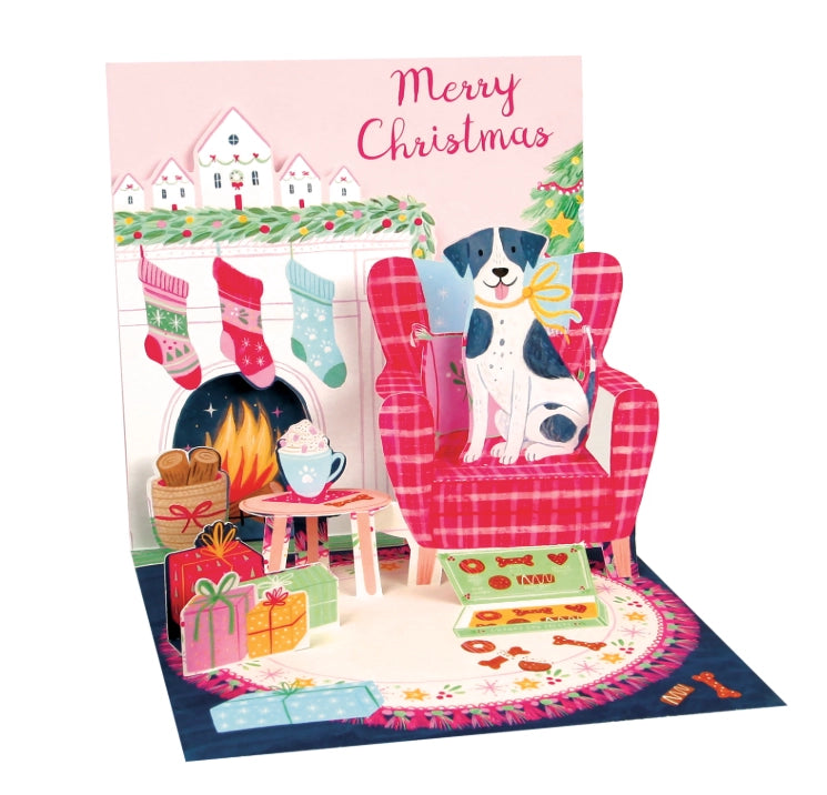 A festive Christmas pop-up card featuring a black and white dog sitting in a red plaid armchair by a fireplace, with stockings, gifts, books, and the words 'Merry Christmas' on a pink background.