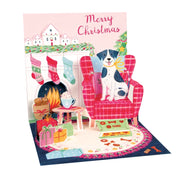 A festive Christmas pop-up card featuring a black and white dog sitting in a red plaid armchair by a fireplace, with stockings, gifts, books, and the words 'Merry Christmas' on a pink background.