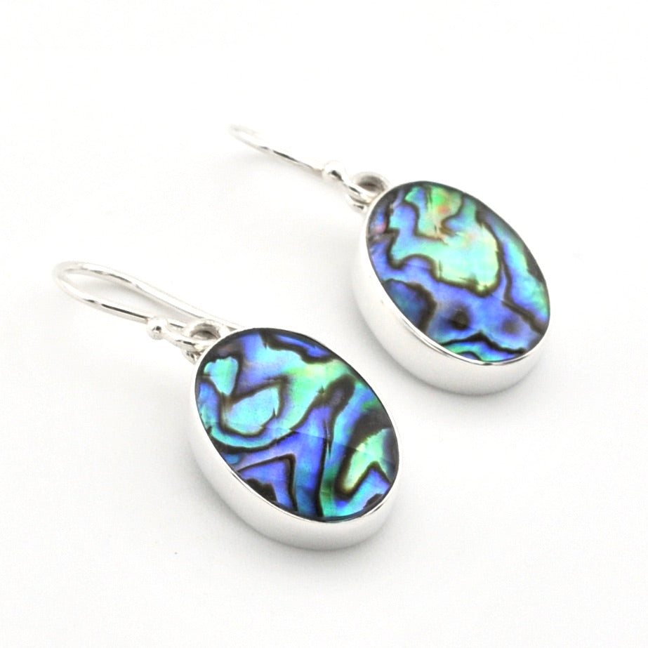 A pair of silver drop earrings featuring oval abalone shell inlays with vibrant blue, green, and purple iridescent patterns, set against a plain white background.