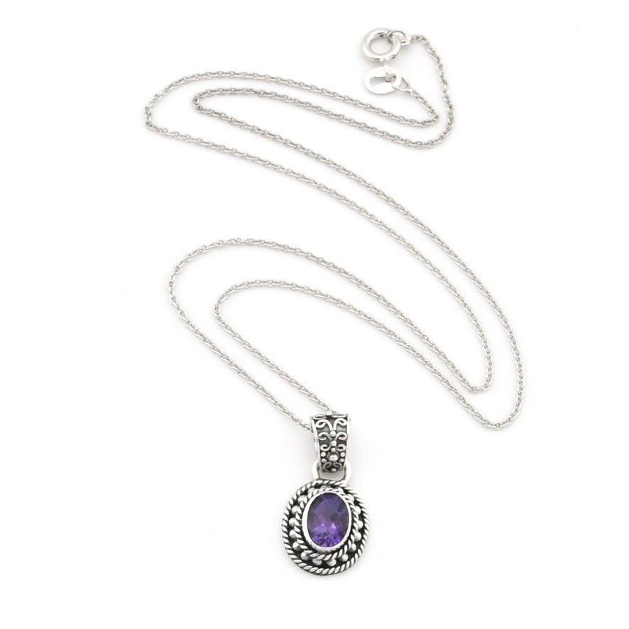 An oval pendant necklace featuring a purple amethyst stone set in a silver-toned, ornate filigree bezel on a delicate silver chain, displayed against a solid white background.