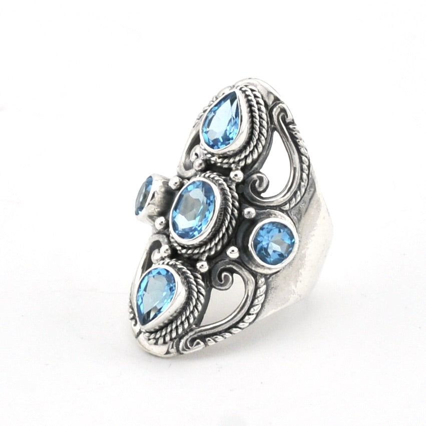 A silver-toned ornate ring featuring five light blue gemstones in a Bali style filigree design, set against a plain white background.