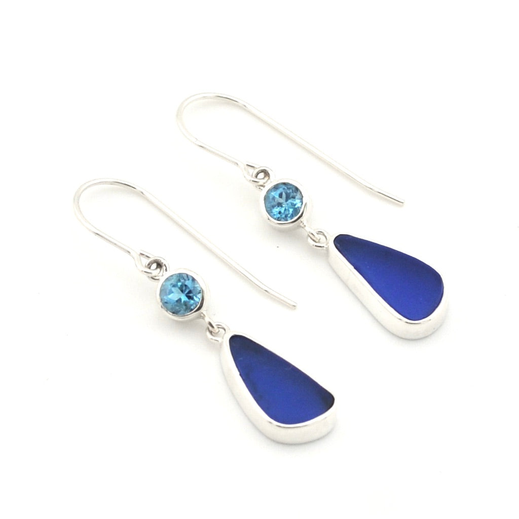 A pair of silver drop earrings featuring a small round light blue topaz and a larger teardrop-shaped dark blue sea glass, shown on a plain white background.