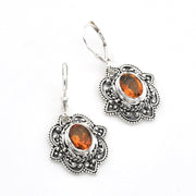 A pair of silver-toned dangle earrings with ornate filigree detailing and oval amber-orange gemstones, shown on a plain white background.
