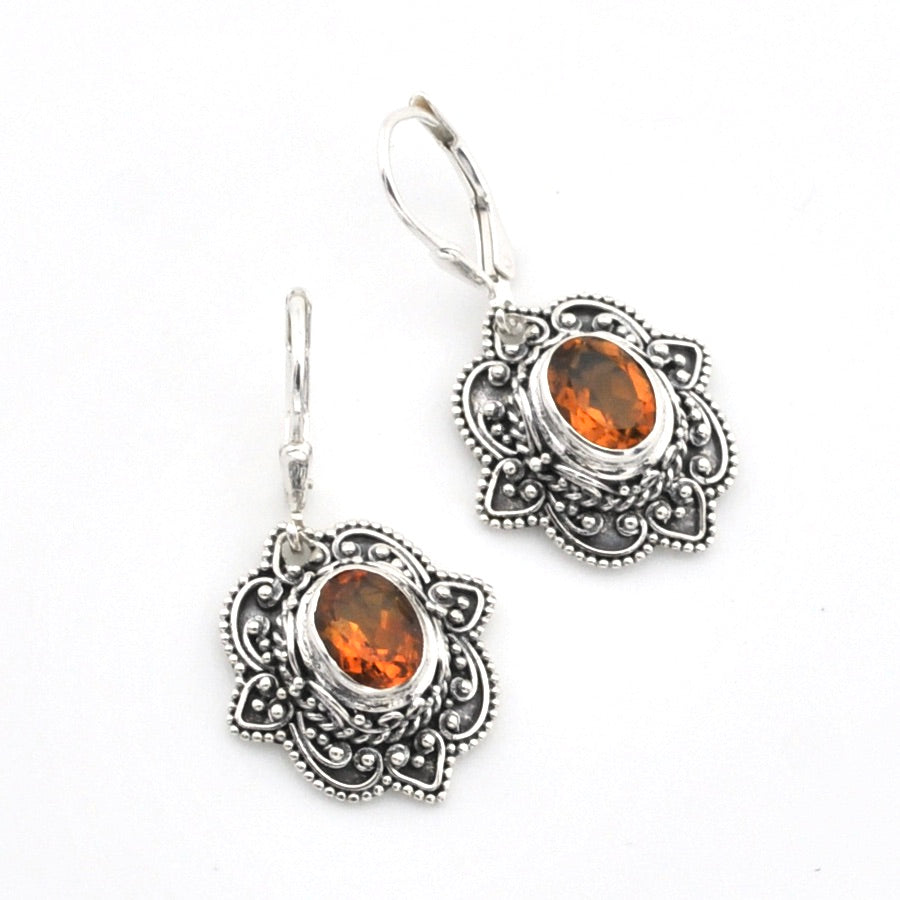 A pair of silver-toned dangle earrings with ornate filigree detailing and oval amber-orange gemstones, shown on a plain white background.