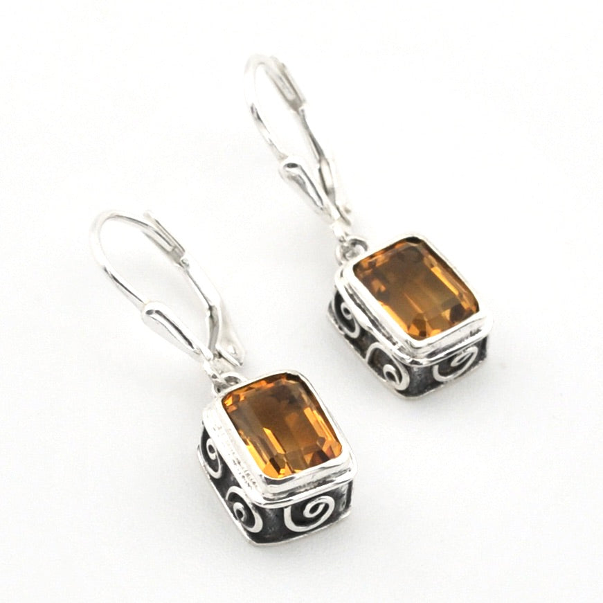 A pair of drop earrings featuring rectangular amber-brown gemstones in silver-toned bezel settings with black and silver spiral engravings, suspended from silver leverback hooks on a white background.