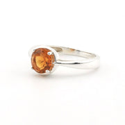 A silver ring featuring a round, faceted orange gemstone in a prong setting, shown on a plain white background.