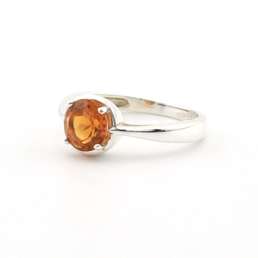 A silver ring featuring a round, faceted orange gemstone in a prong setting, shown on a plain white background.