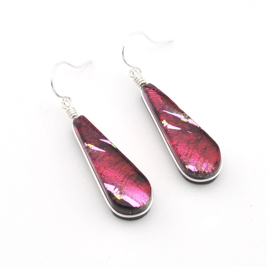 A pair of teardrop-shaped dangle earrings with metallic silver wire-wrapped hooks and bezels, featuring a glossy, textured deep magenta-pink glass inlay with small iridescent flecks, set against a plain white background.