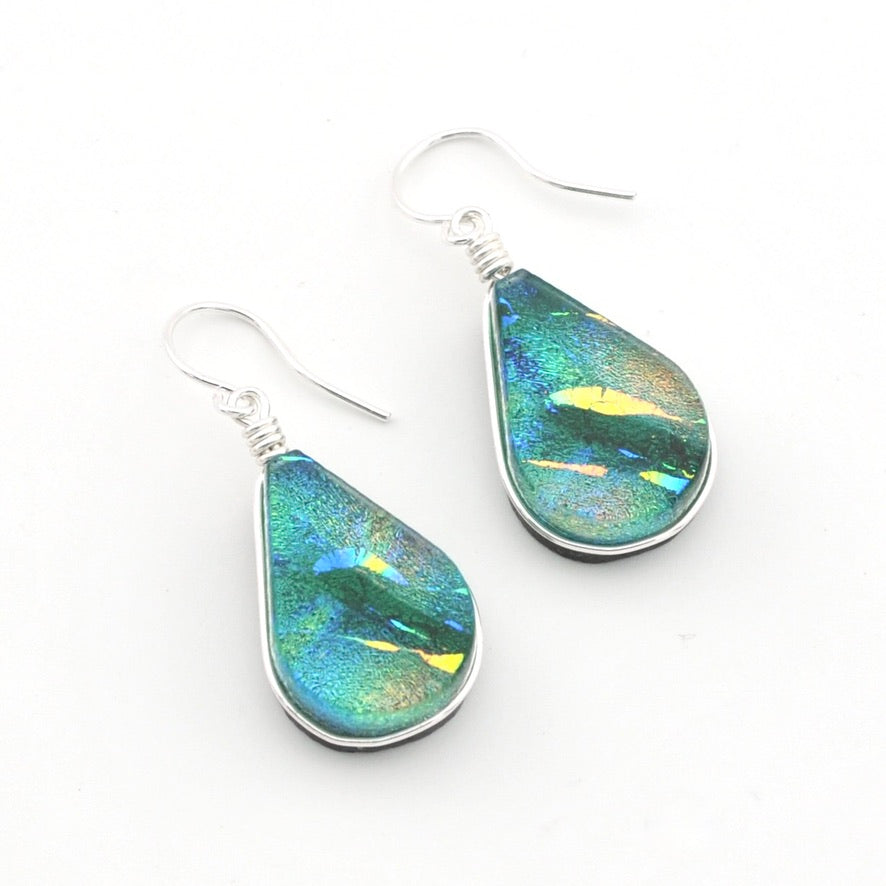 A pair of teardrop-shaped dangle earrings with iridescent blue, green, and gold dichroic glass set in silver, hanging from silver fishhook wires on a plain white background.