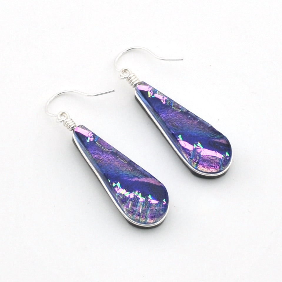 A pair of teardrop-shaped dangle earrings with silver fishhook wires and iridescent purple, blue, and pink glass inlays, displayed on a clean white background.