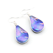 A pair of teardrop-shaped dangle earrings with iridescent blue, purple, and pink dichroic glass pendants in silver bezel settings and silver fishhook ear wires, shown on a plain white background.