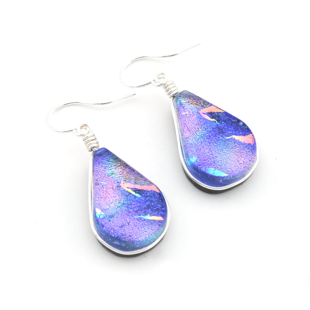 A pair of teardrop-shaped dangle earrings with iridescent blue, purple, and pink dichroic glass pendants in silver bezel settings and silver fishhook ear wires, shown on a plain white background.