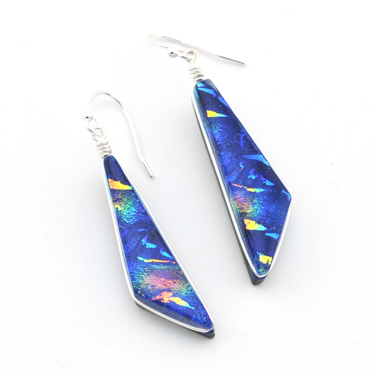A pair of silver wire-wrapped drop earrings featuring elongated, faceted blue iridescent dichroic glass with shimmering yellow, orange, and green flashes, displayed on a clean white background.
