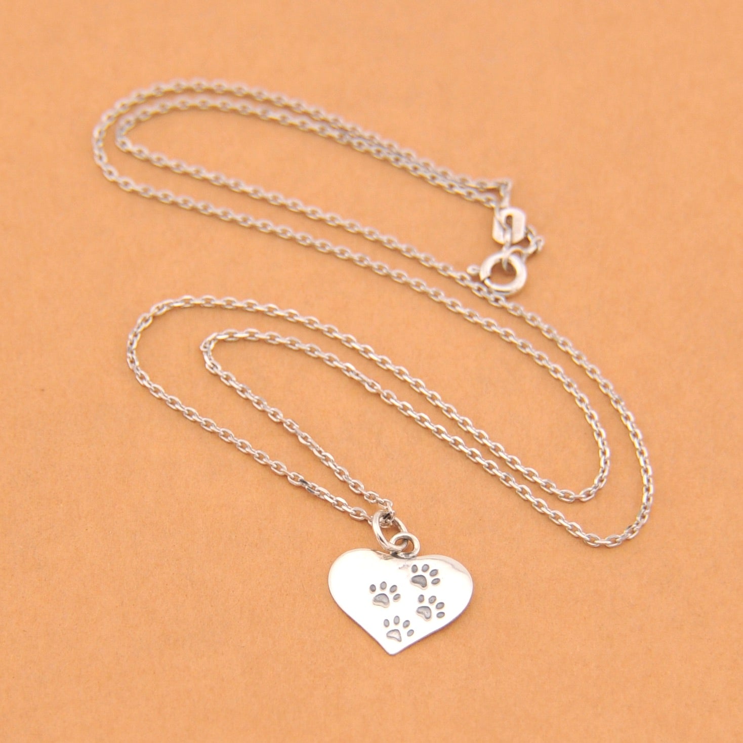 Silver necklace with a heart-shaped pendant featuring paw prints on a beige background