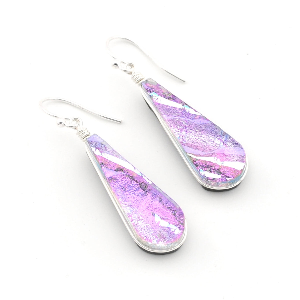 A pair of teardrop-shaped dangling earrings with silver-tone metal frames and hooks, featuring a dichroic glass in shades of purple, pink, and iridescent silver, set against a plain white background.
