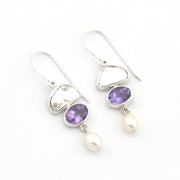 A pair of silver drop earrings featuring oval purple amethyst gemstones and dangling white freshwater pearls, set against a plain white background.