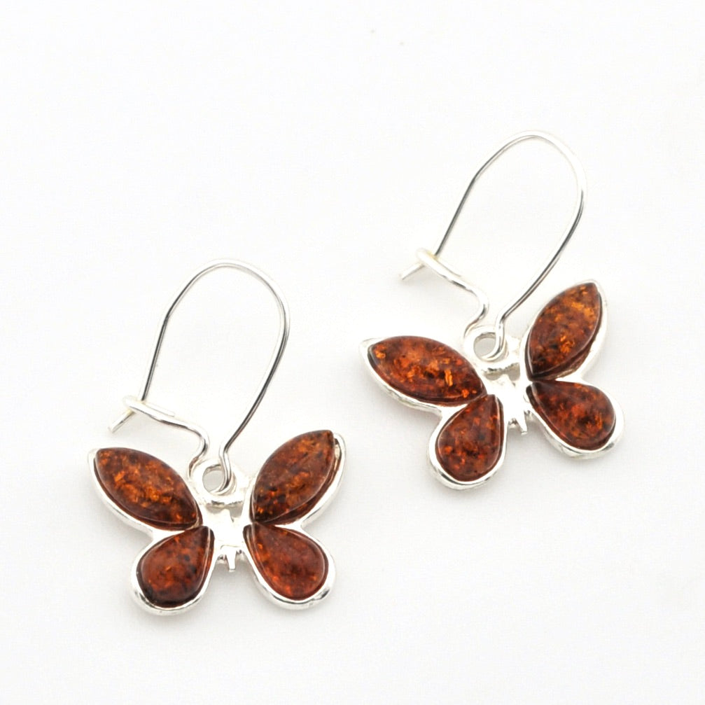 A pair of butterfly-shaped drop earrings featuring polished amber-brown stones in silver settings, suspended from silver fishhook wires on a plain white background.