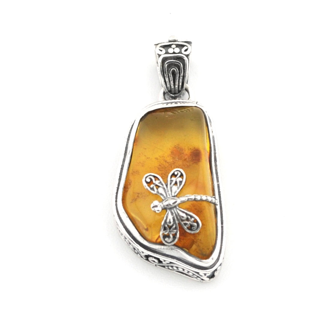 A silver dragonfly pendant set on a polished, irregular-shaped amber stone, shown against a plain white background.
