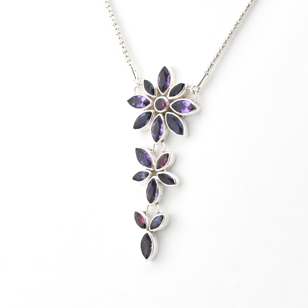 A silver pendant necklace featuring three vertically stacked flower clusters with marquise-cut purple gemstones, shown against a plain white background.