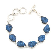 A silver-tone bracelet featuring seven teardrop-shaped blue glitter druse stones set in silver bezels, connected by silver rings and a toggle clasp, shown on a plain white background.