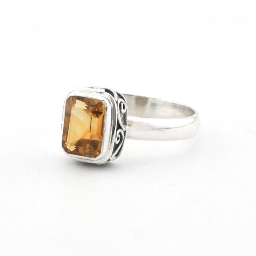 A silver ring featuring a rectangular amber-yellow gemstone with decorative black oxidized scrollwork on the band, set against a plain white background.