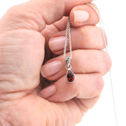 A close-up of a hand holding a silver chain necklace with a teardrop-shaped red gemstone pendant against a white background.