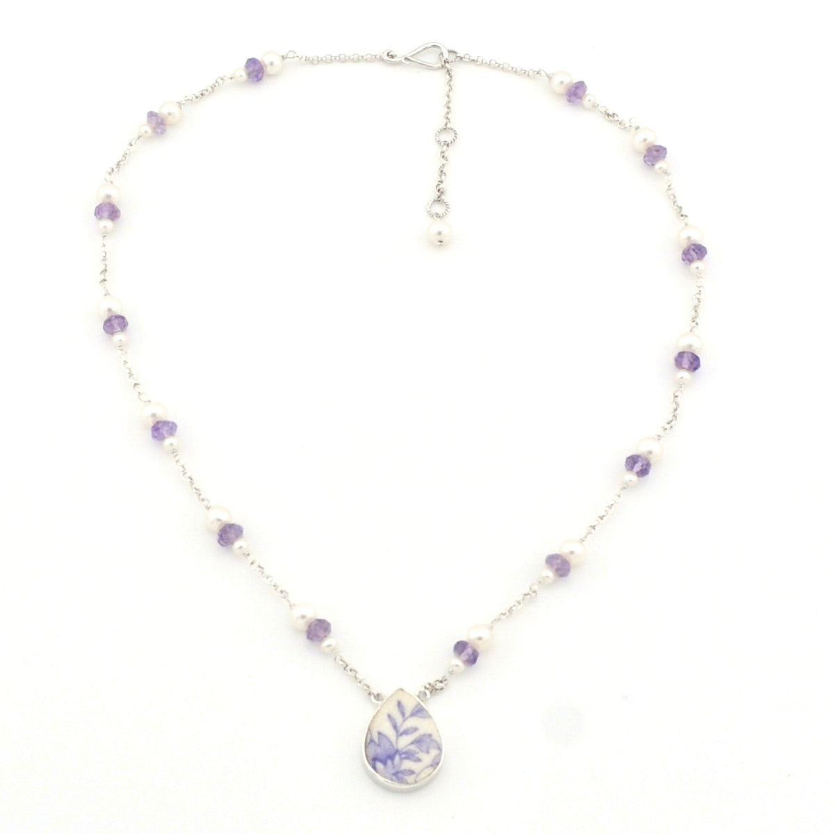 A delicate silver-toned beaded necklace with small purple and white beads, featuring a teardrop-shaped pendant with a purple floral pattern on a white background, set against a plain white backdrop.