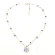 A delicate silver-toned beaded necklace with small purple and white beads, featuring a teardrop-shaped pendant with a purple floral pattern on a white background, set against a plain white backdrop.